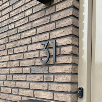 Le numéro de maison Milano Black 15cm de yessential.fr est monté sur un mur de briques beiges près d'une porte de couleur claire, avec une sonnette partiellement visible sur le bord droit.