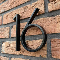 Les numéros de maison Amato Black 12,5 cm de yessential.fr sont montés sur des murs en briques rouges et légèrement éloignés pour projeter une ombre subtile pour un look noir moderne.