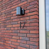 Une applique murale noire moderne et des numéros de maison yessential.fr Amato Black de 15 cm, dont le numéro 17, se dressent sur un mur de briques rouges à côté d'une fenêtre à cadre blanc.