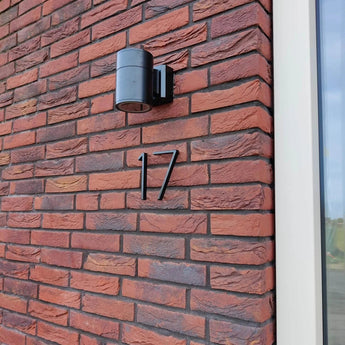 Une applique murale noire moderne et des numéros de maison yessential.fr Amato Black de 15 cm, dont le numéro 17, se dressent sur un mur de briques rouges à côté d'une fenêtre à cadre blanc.