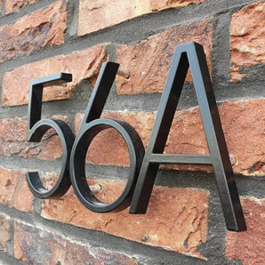 Les numéros de maison Amato Black 12,5 cm de yessential.fr sont montés sur un mur de briques rouges dans un style moderne sans empattement, avec les chiffres et les lettres en relief projetant des ombres saisissantes derrière eux.