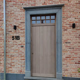Une porte d'entrée en bois clair avec des fenêtres rectangulaires au-dessus, placées dans un cadre en pierre grise. Le mur de briques rouge clair comporte un numéro de maison yessential.fr Adamo Black de 15 cm « 51B » sur la gauche et deux appliques noires flanquent la porte.