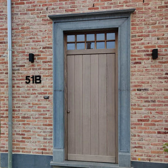 Une porte d'entrée en bois clair avec des fenêtres rectangulaires au-dessus, placées dans un cadre en pierre grise. Le mur de briques rouge clair comporte un numéro de maison yessential.fr Adamo Black de 15 cm « 51B » sur la gauche et deux appliques noires flanquent la porte.