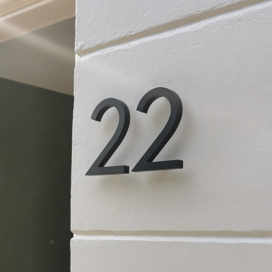 Le numéro de maison Milano Black 10 cm de yessential.fr présente un élégant « 22 » noir sur un mur blanc lisse. Fabriqué en aluminium, il crée des ombres douces, rendant ce numéro de maison à la fois fonctionnel et élégant.