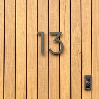 Une porte en bois avec des panneaux verticaux affiche le numéro de maison « 13 » yessential.fr Milano Black 15 cm en métal moderne, avec une sonnette ou un interphone noir monté à droite.