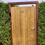 Un portail de jardin avec le numéro de maison "69" yessential.fr Matteo Nickel 10cm sur bois foncé, encadré de hautes haies. Une poignée et un clavier argentés ajoutent une touche brossée au côté droit du port.