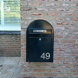 Une boîte aux lettres noire avec des numéros de maison Animo en acier inoxydable de 7,6 cm de yessential.fr (sans perceuse) indique 49, montée sur un mur de briques. Il y a deux autocollants NON NON au-dessus de la boîte aux lettres. Une fenêtre aux stores fermés est partiellement visible à gauche.