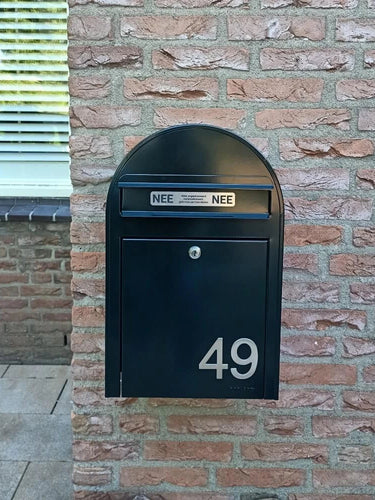 Une boîte aux lettres noire avec des numéros de maison Animo en acier inoxydable de 7,6 cm de yessential.fr (sans perceuse) indique 49, montée sur un mur de briques. Il y a deux autocollants NON NON au-dessus de la boîte aux lettres. Une fenêtre aux stores fermés est partiellement visible à gauche.