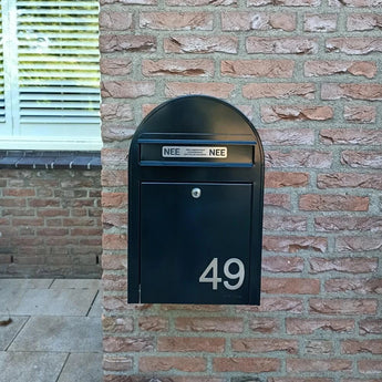Une boîte aux lettres noire avec des numéros de maison Animo en acier inoxydable de 7,6 cm de yessential.fr (sans perceuse) indique 49, montée sur un mur de briques. Il y a deux autocollants NON NON au-dessus de la boîte aux lettres. Une fenêtre aux stores fermés est partiellement visible à gauche.