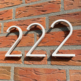 Les numéros de maison Amato White de 12,5 cm de yessential.fr en « blanc 222 » sont montés sur un mur de briques rouges avec du mortier gris, qui projette des ombres saisissantes à la lumière du soleil pour un look moderne.