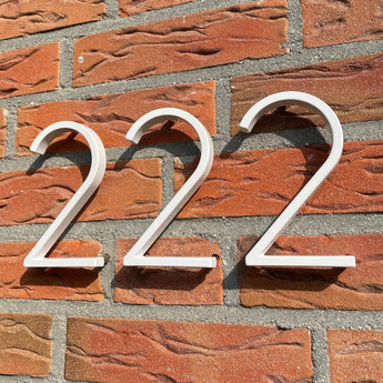 Les numéros de maison Amato White de 12,5 cm de yessential.fr en « blanc 222 » sont montés sur un mur de briques rouges avec du mortier gris, qui projette des ombres saisissantes à la lumière du soleil pour un look moderne.