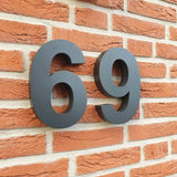 Les Angelo Black 20cm de yessential.fr sont de grands numéros de maison 3D noirs, montés sur un mur de briques rouges avec des joints visibles et une texture rustique, idéaux comme numéros de maison modernes.