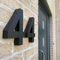 Les numéros de maison Angelo Black 20 cm de yessential.fr sont montés sur un mur de briques claires à côté d'une porte gris foncé moderne avec des inserts en verre rectangulaires.