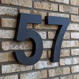 Numéro de maison 57 Angelo Black 30 cm de yessential.fr en style 3D, monté sur un mur de briques beiges - moderne et élégant comme numéro de maison pour n'importe quelle maison ou bâtiment.