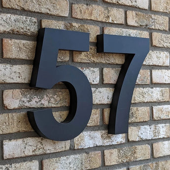 Numéro de maison 57 Angelo Black 30 cm de yessential.fr en style 3D, monté sur un mur de briques beiges - moderne et élégant comme numéro de maison pour n'importe quelle maison ou bâtiment.