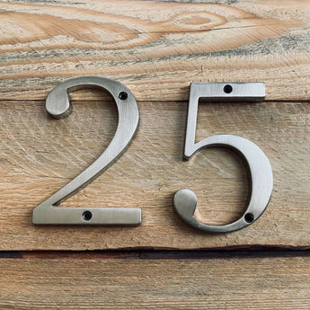 Les numéros de maison Matteo Nickel 10 cm de yessential.fr sont placés sur du bois clair avec un grain de bois visible et des trous de vis dans chaque numéro.