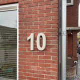 Le béton Enrico 30 cm de yessential.fr est un grand numéro de maison 3D blanc « 10 » affiché sur un mur extérieur en brique rouge à côté d'une fenêtre avec des stores fermés et un tuyau d'évacuation vertical argenté.
