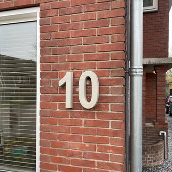 Le béton Enrico 30 cm de yessential.fr est un grand numéro de maison 3D blanc « 10 » affiché sur un mur extérieur en brique rouge à côté d'une fenêtre avec des stores fermés et un tuyau d'évacuation vertical argenté.