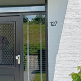 Entrée de maison avec une porte noire avec panneau grillagé et poignée blanche ; Un numéro de maison Animo Black 7,6 cm (sans perçage) de yessential.fr est monté sur le mur de briques blanches. Une colline herbeuse et une clôture sont visibles à travers la vitre.