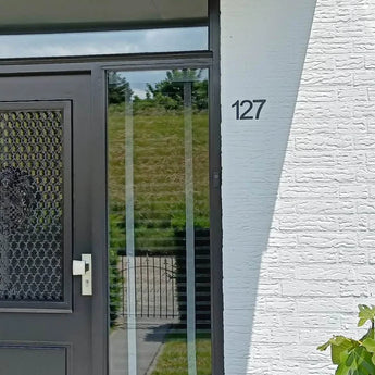 Entrée de maison avec une porte noire avec panneau grillagé et poignée blanche ; Un numéro de maison Animo Black 7,6 cm (sans perçage) de yessential.fr est monté sur le mur de briques blanches. Une colline herbeuse et une clôture sont visibles à travers la vitre.