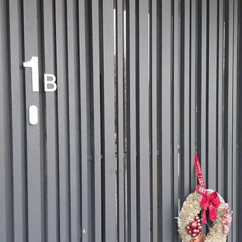 Une porte à lattes verticales sombres étiquetée « 1B » avec une couronne festive d'ornements et de rubans rouges, avec le numéro de maison Animo de 15 cm en acier inoxydable yessential.fr (sans perçage) sur la droite pour un look élégant et moderne.