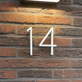 Monté sur un mur de briques rouges et marron foncé avec une lampe rectangulaire au-dessus, le numéro de maison Amato White 12,5 cm de yessential.fr offre un style moderne - idéal pour tous ceux qui recherchent des numéros de maison modernes et saisissants pour leur maison.