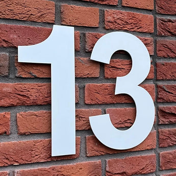 L'Adamo White 30 cm de yessential.fr est un grand numéro 13 blanc, conçu dans le style Adamo White et monté sur un mur de briques rouges - un numéro de maison frappant en acier inoxydable pour un adressage clair.