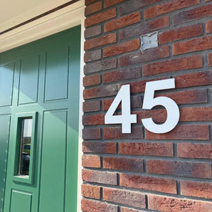 Un gros plan d'un mur de briques montre le numéro de maison « 45 » yessential.fr Adamo White de 15 cm à côté d'une porte d'entrée verte avec une fenêtre rectangulaire, la lumière du soleil projetant des ombres sur les deux.