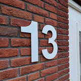 Le numéro de maison Adamo White 30 cm de yessential.fr, en taille 13, est monté sur un mur de briques rouges à côté d'une fenêtre ou d'une porte à cadre blanc.