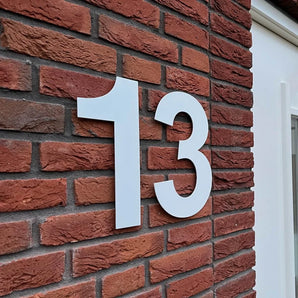 Le numéro de maison Adamo White 30 cm de yessential.fr, en taille 13, est monté sur un mur de briques rouges à côté d'une fenêtre ou d'une porte à cadre blanc.