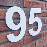 Le numéro de maison 95 Adamo White 20 cm de yessential.fr se détache magnifiquement sur un mur de briques rouges, rendant les numéros de bâtiment ou de maison clairement visibles.