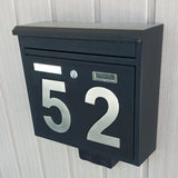 Une boîte aux lettres en métal noir avec de grands numéros de maison Animo en acier inoxydable de 15 cm de yessential.fr est montée sur un mur léger à panneaux verticaux. La boîte aux lettres dispose d'une fente pour le courrier, d'un trou de serrure et d'un support de plaque signalétique rectangulaire.