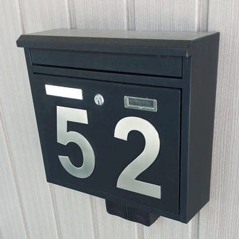 Une boîte aux lettres en métal noir avec de grands numéros de maison Animo en acier inoxydable de 15 cm de yessential.fr est montée sur un mur léger à panneaux verticaux. La boîte aux lettres dispose d'une fente pour le courrier, d'un trou de serrure et d'un support de plaque signalétique rectangulaire.