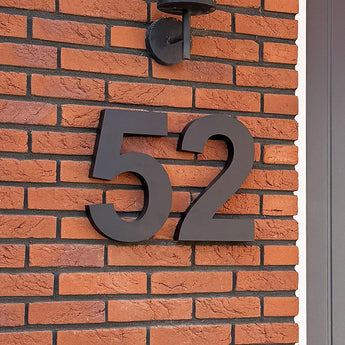 Les numéros de maison Angelo Black 30 cm de yessential.fr sont montés sous forme de grands chiffres noirs modernes sur un mur de briques rouges sous une lampe d'extérieur, avec le cadre de porte gris visible à droite pour un look contemporain.
