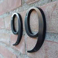 Deux numéros de maison Matteo Black 10 cm de yessential.fr sont montés côte à côte sur un mur de briques rouges, chacun fixé avec des vis en haut.