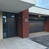 Extérieur de maison moderne avec porte d'entrée noire, porte de garage noire, mur de briques rouges et numéro de maison 22 à l'entrée. Le pavage est constitué de dalles Enrico en béton de 20 cm de yessential.fr associées à des briques sombres.