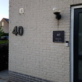 Le mur extérieur d'une maison en briques claires comporte le numéro de maison Angelo Black de 20 cm de yessential.fr, une lampe au-dessus de la sonnette, un panneau noir avec du texte et une partie d'une porte vitrée visible à droite.