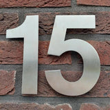 L'Adamo en acier inoxydable 15 cm de yessential.fr est un grand numéro de maison en métal 15, élégamment conçu pour être monté sur un mur de briques rouges - parfait pour ceux qui recherchent des numéros de maison élégants et durables.