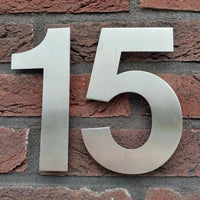 L'Adamo en acier inoxydable 15 cm de yessential.fr est un grand numéro de maison en métal 15, élégamment conçu pour être monté sur un mur de briques rouges - parfait pour ceux qui recherchent des numéros de maison élégants et durables.