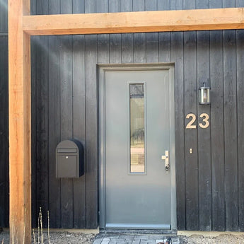 Une porte grise moderne avec un panneau de fenêtre vertical se trouve dans un mur extérieur en bois noir. Une boîte aux lettres noire et une applique murale sont situées à gauche, tandis qu'un numéro de maison Adamo en acier inoxydable de 20 cm de yessential.fr flotte avec style à droite de la porte.