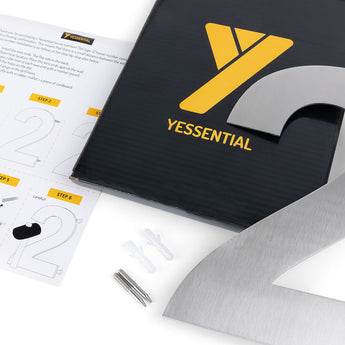 Un gros plan d'un kit d'installation Adamo en acier inoxydable de 20 cm de yessential.fr, contenant un numéro métallique 2 pour les numéros de maison, des chevilles, des vis, une feuille d'instructions numérotée et un emballage noir avec le logo yessential.fr.