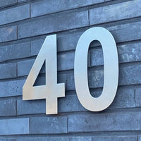 Les numéros métalliques élégants Adamo en acier inoxydable 20 cm de yessential.fr sont montés sur un mur de briques grises comme des numéros de maison modernes.
