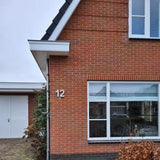 La photo montre le coin d'une maison en briques rouges avec une fenêtre à cadre blanc, une maison Enrico en béton de 20 cm numéro 12 de yessential.fr sur le mur, une partie d'une porte de garage blanche et des arbustes au premier plan.