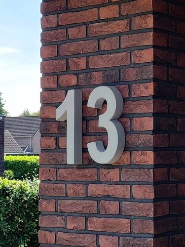 Le béton Enrico 30 cm de yessential.fr est un chiffre 13 blanc audacieux dans le style d'un numéro de maison 3D, monté sur une colonne de briques rouges et marron foncé à l'extérieur d'un bâtiment, avec de la verdure et des maisons voisines en arrière-plan.