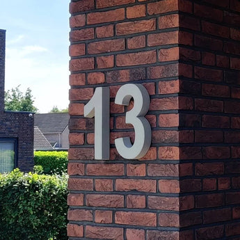 Le béton Enrico 30 cm de yessential.fr est un chiffre 13 blanc audacieux dans le style d'un numéro de maison 3D, monté sur une colonne de briques rouges et marron foncé à l'extérieur d'un bâtiment, avec de la verdure et des maisons voisines en arrière-plan.
