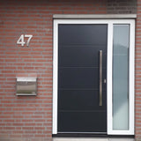 Porte d'entrée noire moderne avec une haute poignée argentée, un cadre blanc et un panneau vertical en verre dépoli à droite. A gauche, un numéro de maison Adamo en acier inoxydable de 20 cm de yessential.fr et une boîte aux lettres en acier inoxydable sont montés sur le mur de briques rouges.