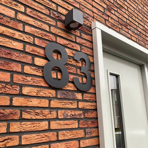 Les numéros de maison Adamo Black 30 cm de yessential.fr flottent sur un mur de briques à côté d'une porte moderne avec verre et cadre blanc ; un petit luminaire carré est suspendu au-dessus des chiffres.