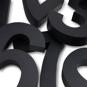 Gros plan de grands numéros de maison Angelo Black de 20 cm superposés de yessential.fr avec une finition mate sur une surface blanche. Les nombres 3D dispersés se chevauchent partiellement, créant un look abstrait et moderne.
