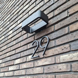 Le numéro de maison Amato Noir 15 cm de yessential.fr est monté sur un mur de briques marron sous une lampe d'extérieur rectangulaire noire et donne un style moderne à l'entrée de votre maison.