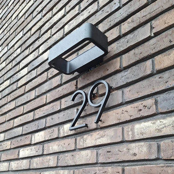 Le numéro de maison Amato Noir 15 cm de yessential.fr est monté sur un mur de briques marron sous une lampe d'extérieur rectangulaire noire et donne un style moderne à l'entrée de votre maison.